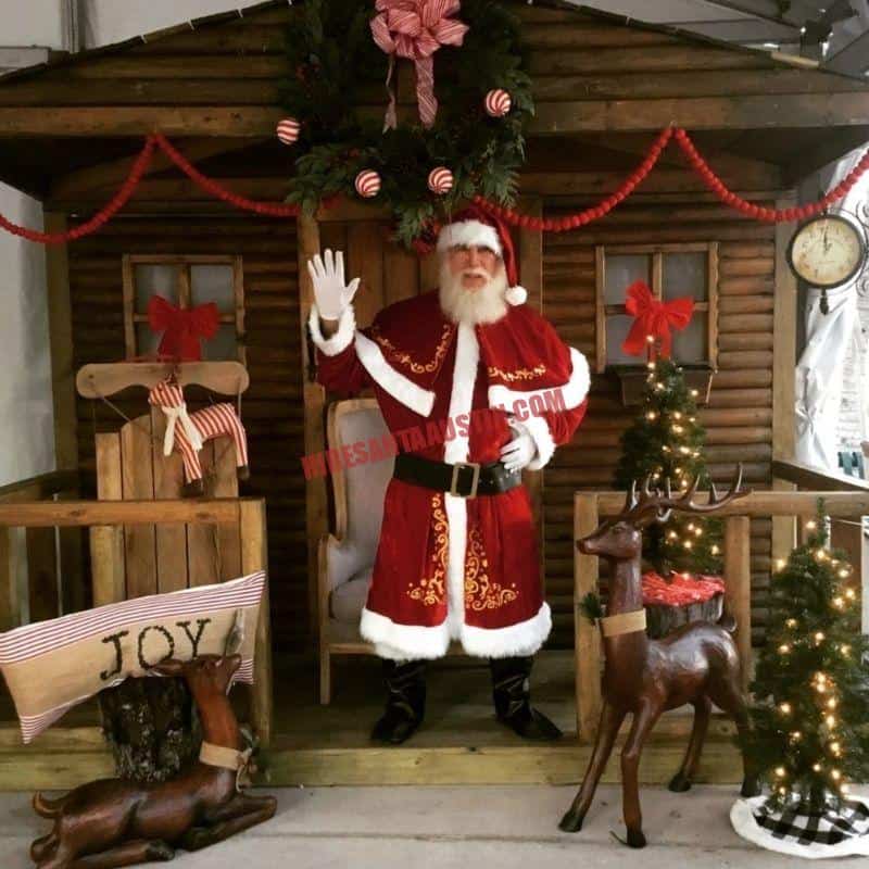 Real bearded Santa in a classic red suit waves from a wooden cabin set with wreaths and reindeer statues.
