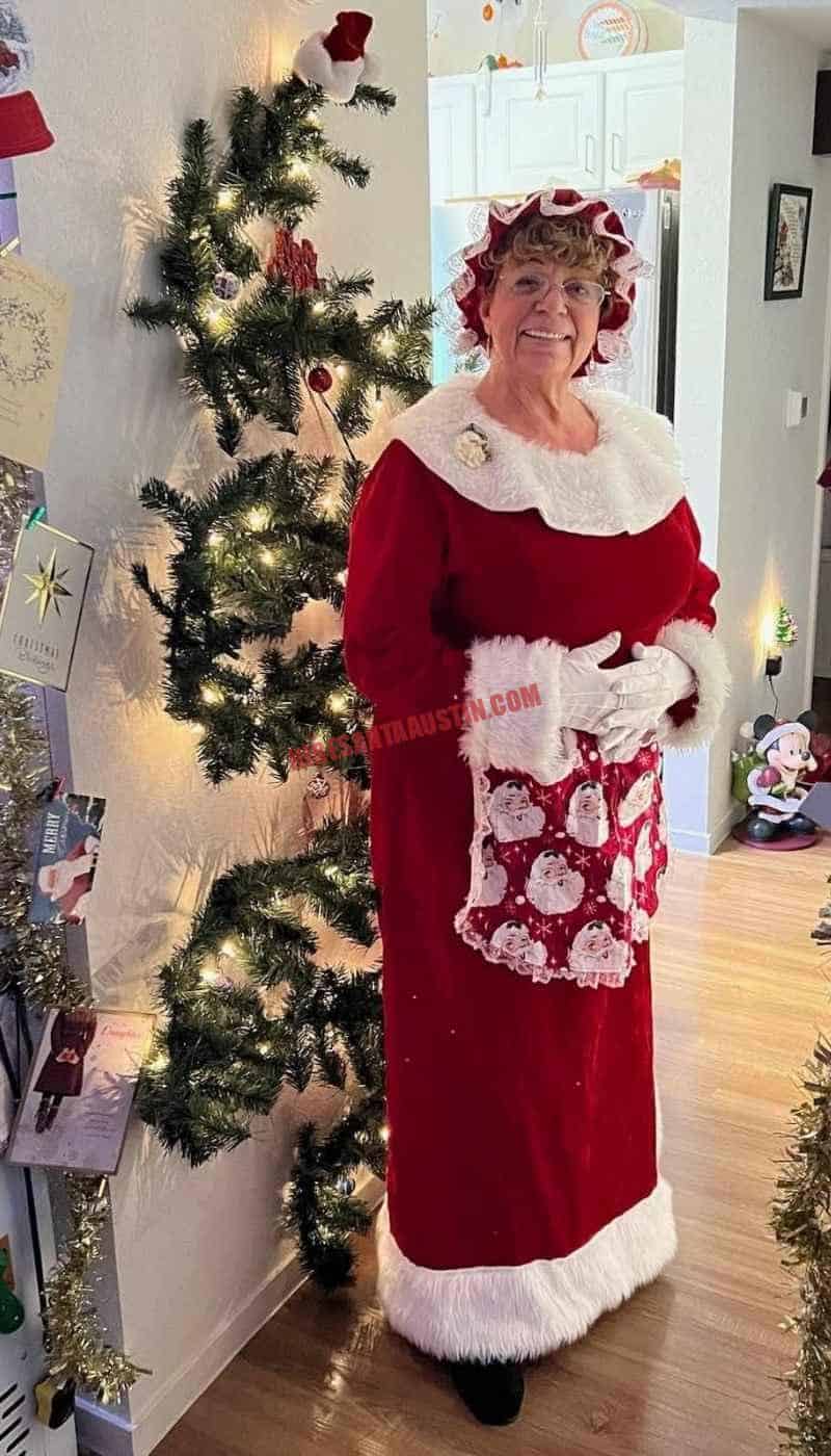 Mrs. Claus for hire in Austin, in a red dress, white apron, and gloves beside a lit tree.