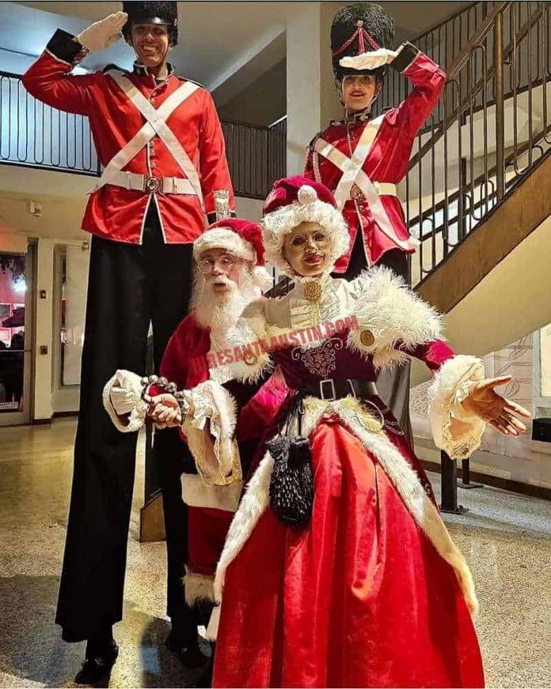 Corporate Santa with Mrs. Claus and two stilt nutcracker soldiers in a decorated lobby.