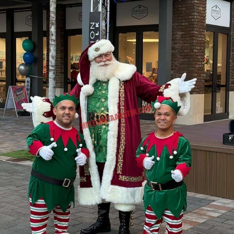 Santa Claus impersonator in a burgundy coat and green vest stands with two elves outside a shop.