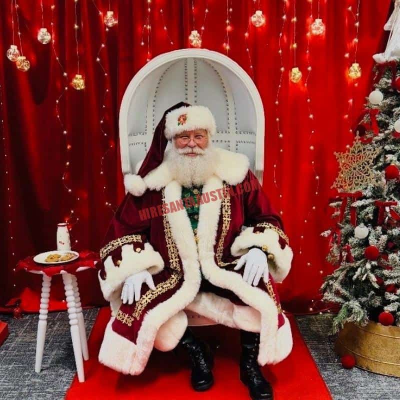 Hire Santa Round Rock: real beard Santa Claus in a burgundy coat sits on a white high-back chair beside cookies and a small tree.